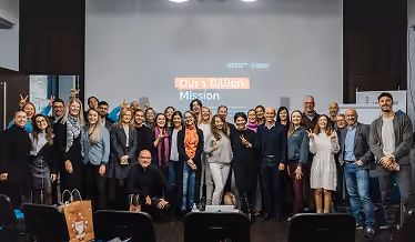Group of diverse people standing and smiling in front of a screen with 'Our 1 Billion Mission' text.