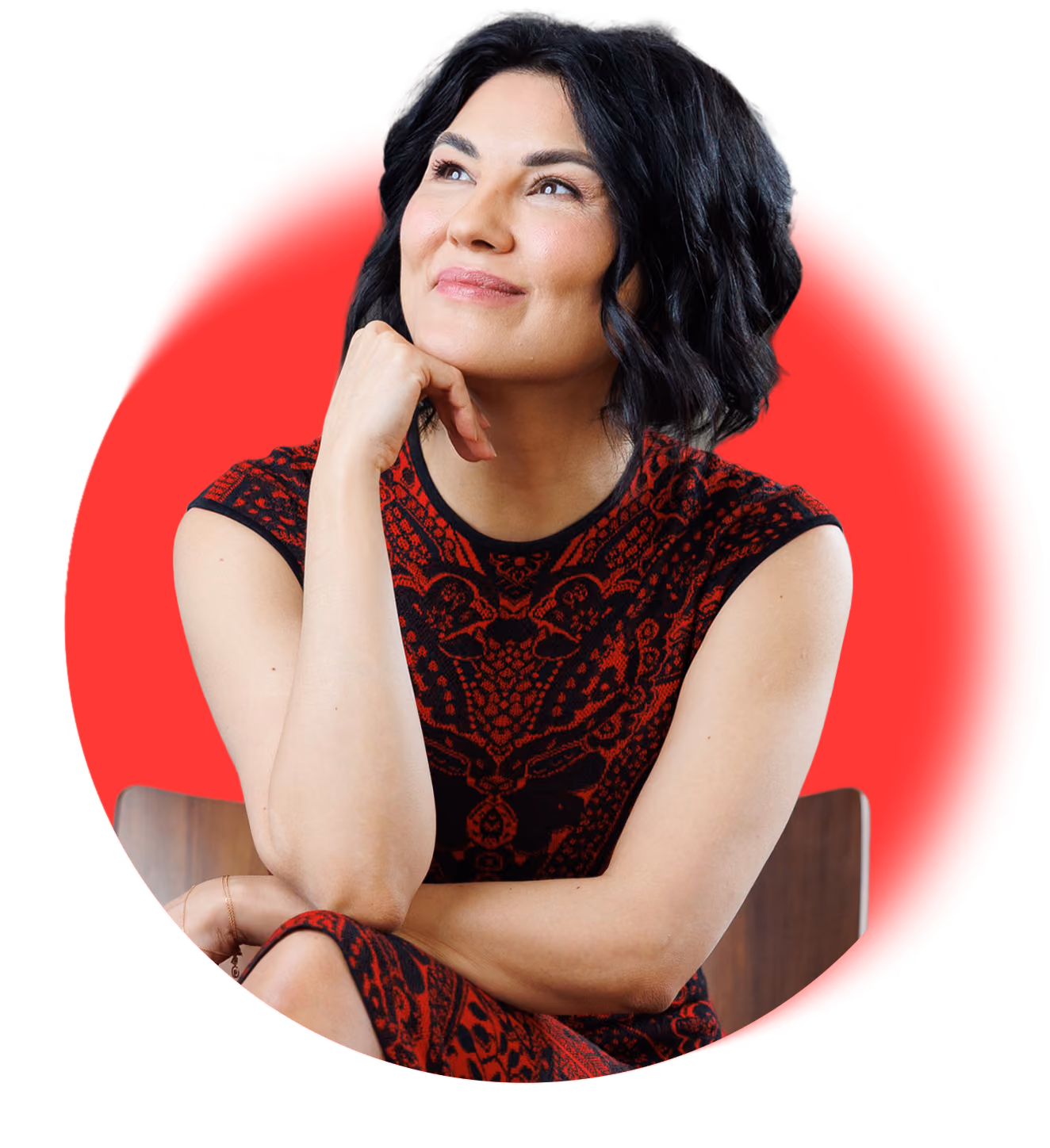 Woman with short black hair wearing a patterned red and black dress, sitting and looking up with a smile.