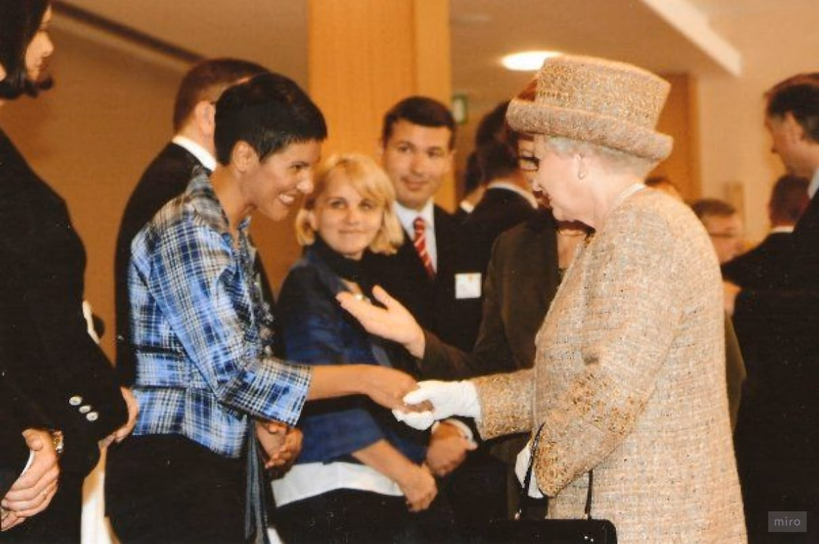 Woman in blue plaid jacket shaking hands with elderly woman in beige suit and hat at formal event.