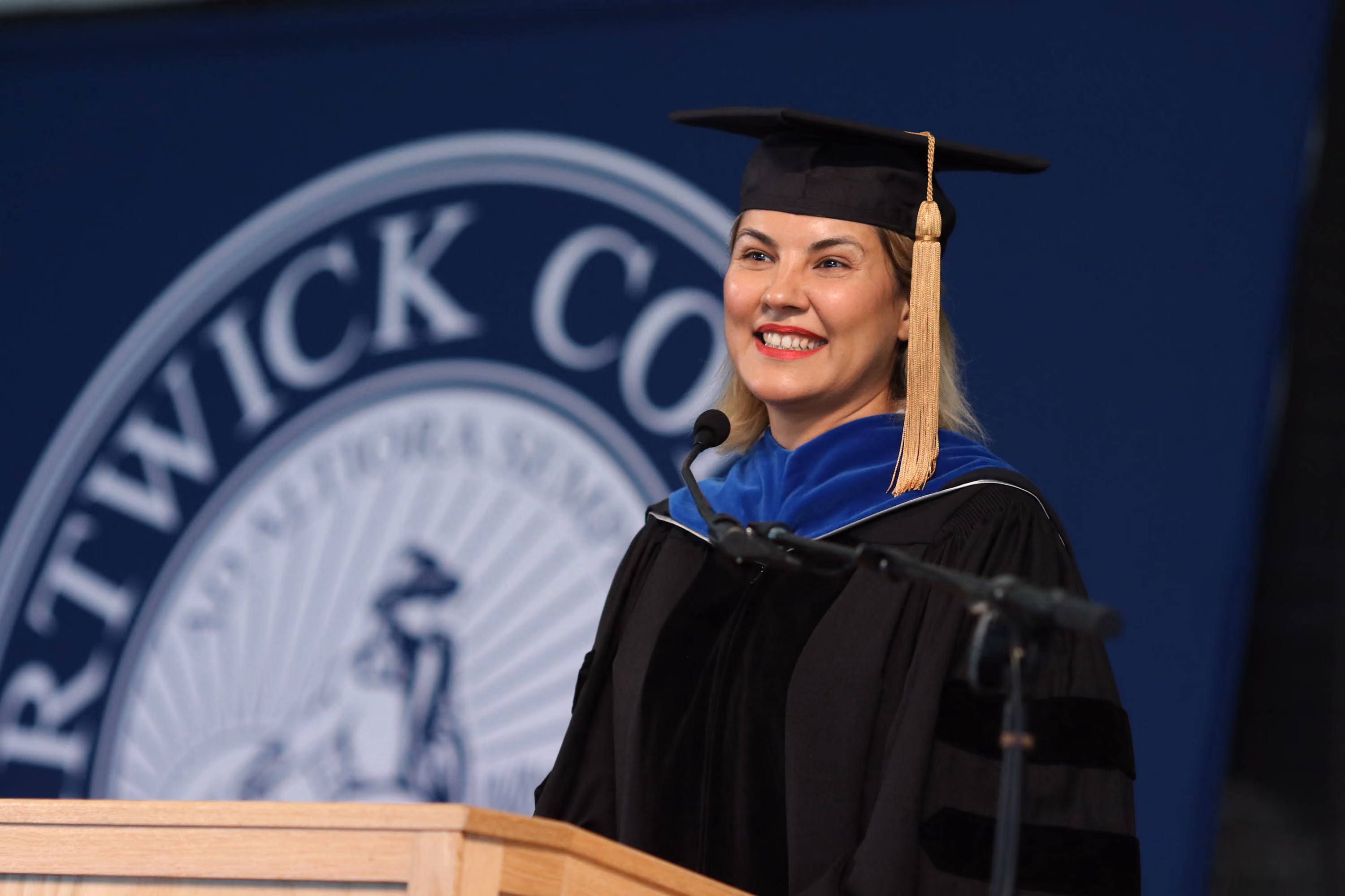 Smiling woman in graduation cap and gown speaking at a podium with a microphone.