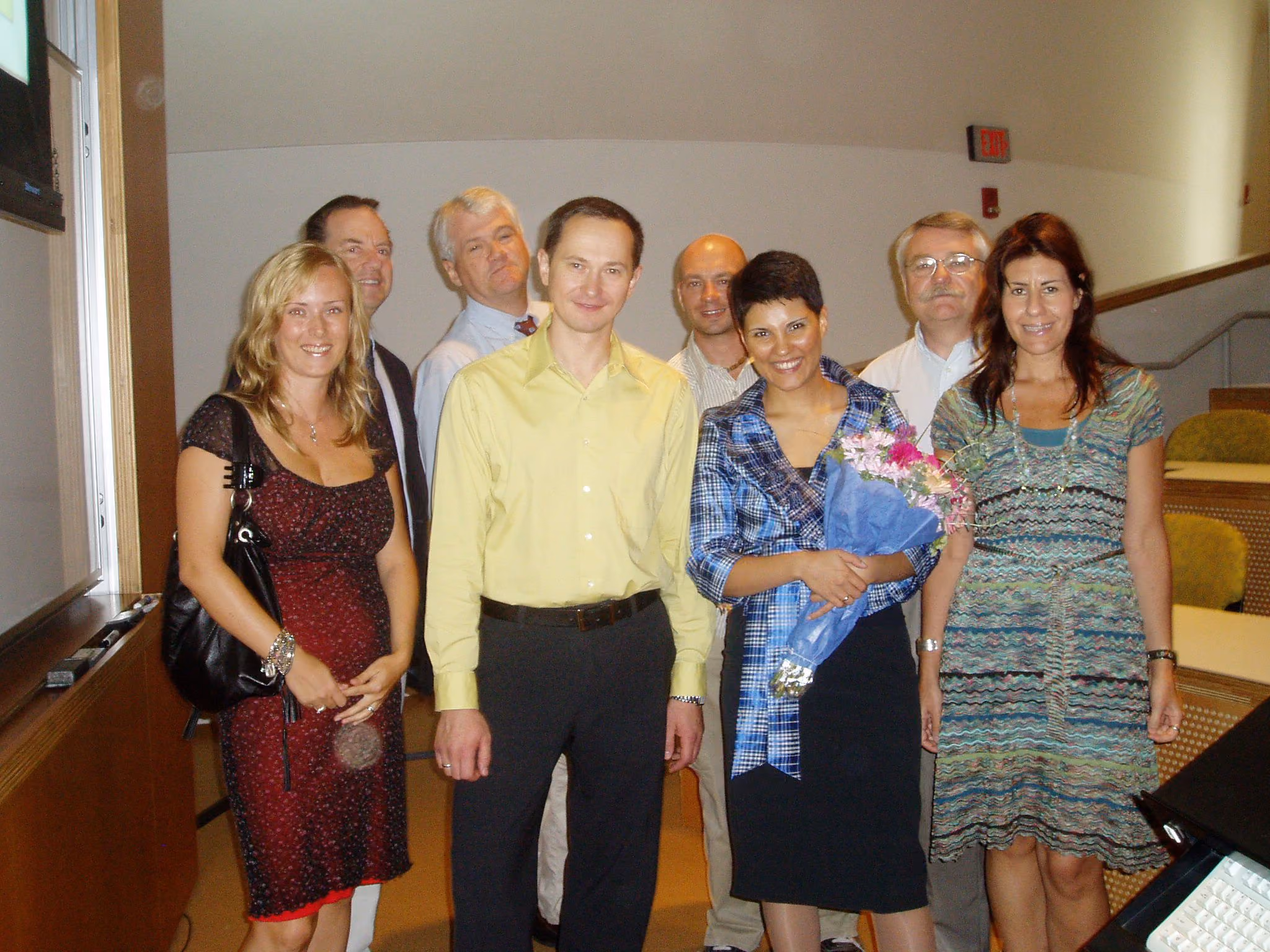 Group of seven adults smiling indoors, one woman holds a bouquet of flowers.