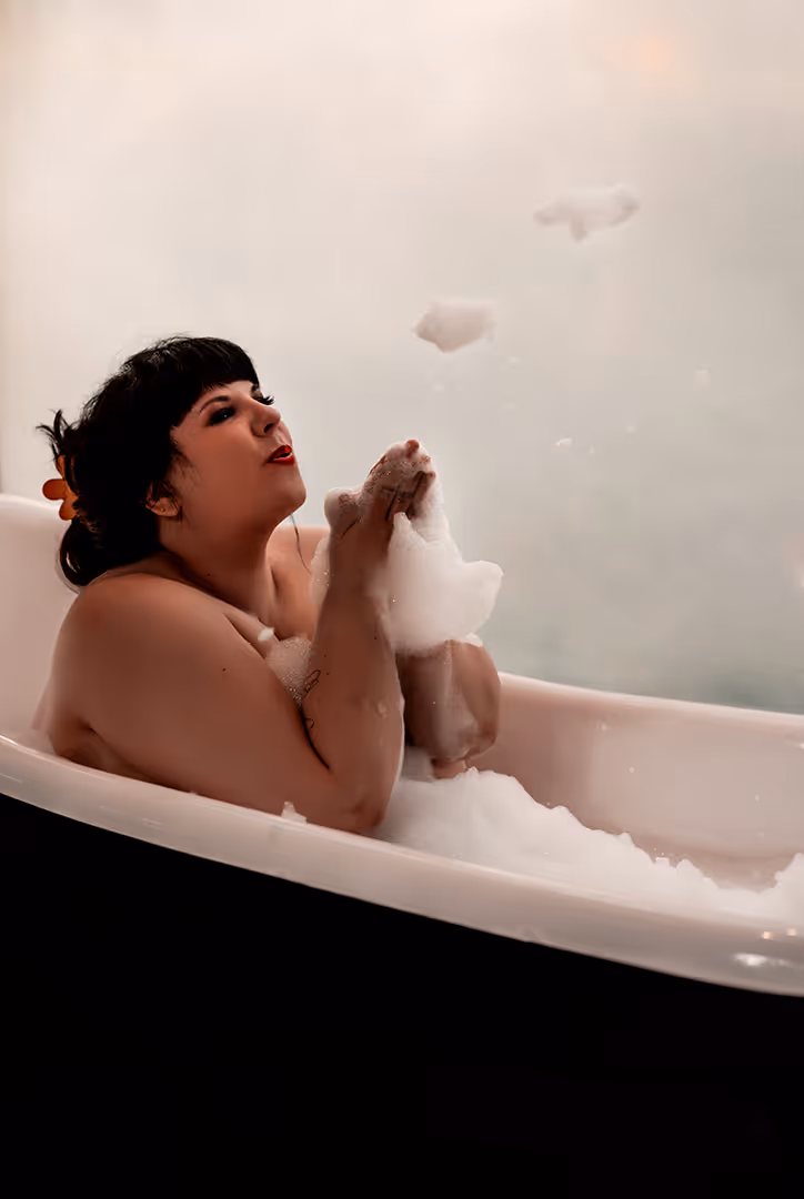 Woman with dark hair blowing bubbles while relaxing in a bathtub filled with foam.
