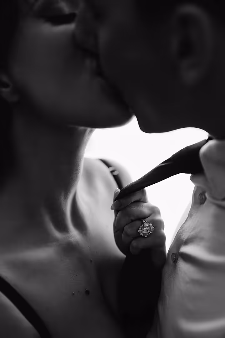 Black and white close-up of a couple kissing, showing a woman's hand with an ornate engagement ring holding the man's tie.