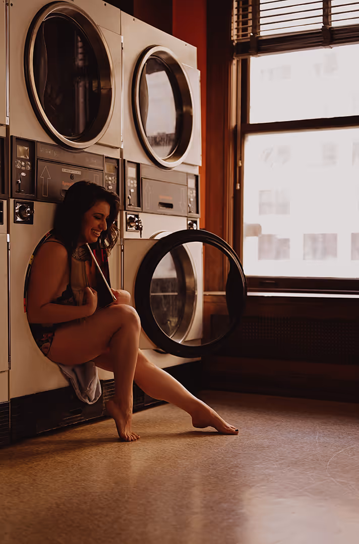 Woman sitting inside an open front-loading washing machine door, smiling and holding a book, in a laundry room with large windows.