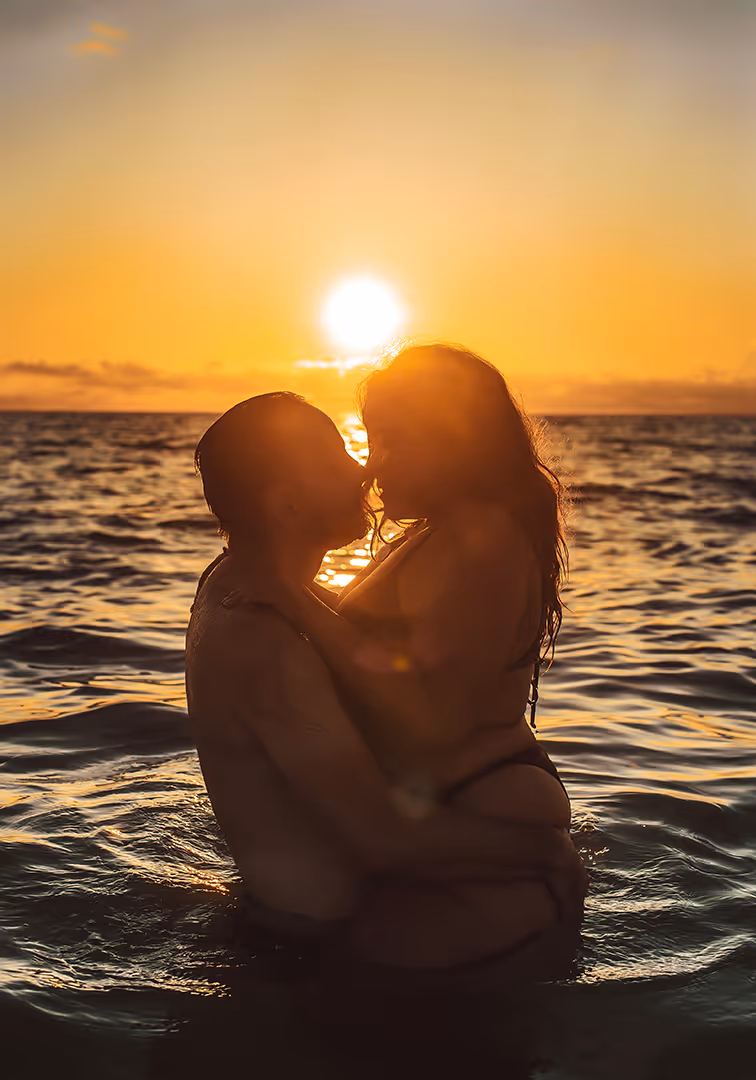 Silhouetted couple embracing and about to kiss while standing in the ocean at sunset.