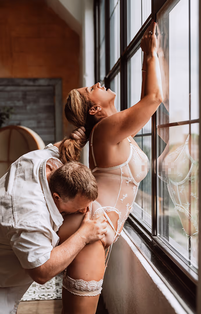 Couple in intimate moment by a large window, woman wearing sheer lingerie standing while man kisses her thigh.