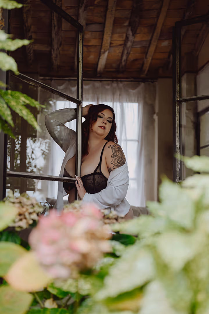 Woman in black lace lingerie and white shirt posing by an open window with a wooden ceiling and blurred greenery in the foreground.