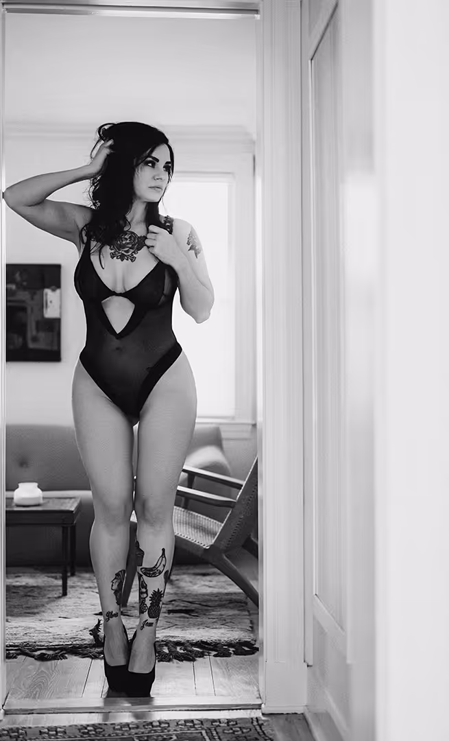 Black-and-white photo of a tattooed woman in a black bodysuit and heels standing indoors near a doorway.