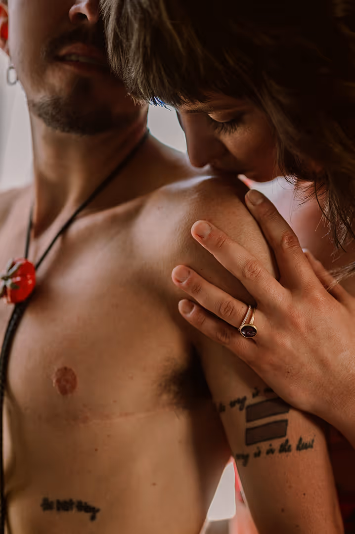 Woman wearing a ring gently resting her hand and kissing a shirtless man's shoulder with visible tattoos.