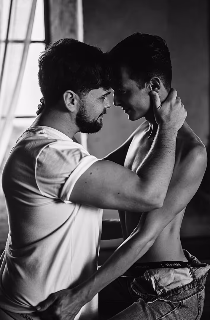 Black and white photo of two men embracing closely, forehead to forehead, one wearing a white t-shirt and the other shirtless with jeans.