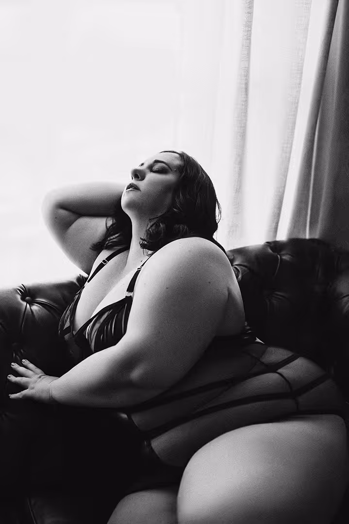 Black and white photo of a plus-size woman reclining on a leather sofa in lingerie, eyes closed and hand behind her head.