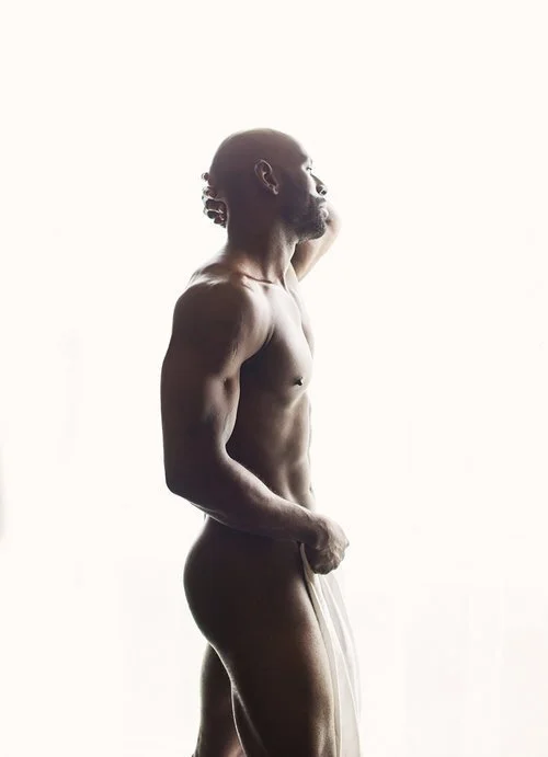 Side profile of a muscular man standing against a bright background, holding a sheer cloth covering his lower body.