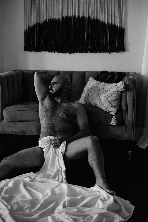 Black and white photo of a shirtless man sitting on the floor leaning against a couch, holding a white sheet and looking to the side.