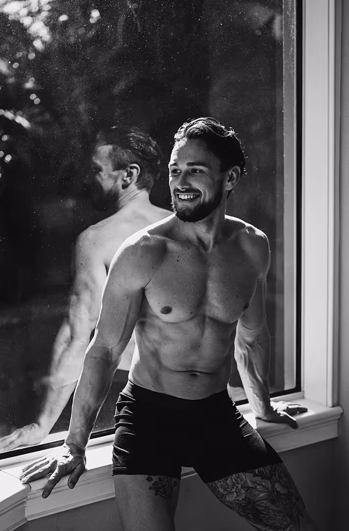 Smiling muscular shirtless man in black shorts sitting on a window ledge with his reflection visible in the glass.