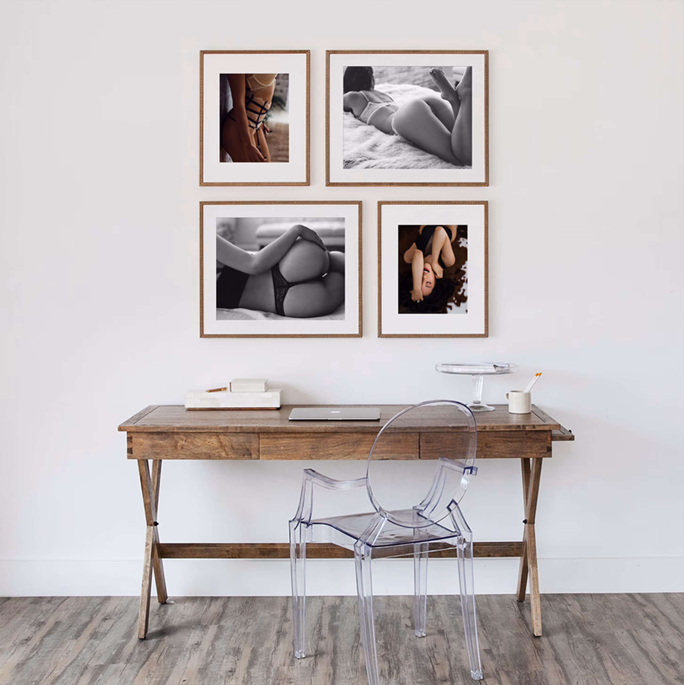 Wooden desk with laptop, books, and cup, in front of a wall with four framed artistic photos of women in lingerie, and a transparent chair.