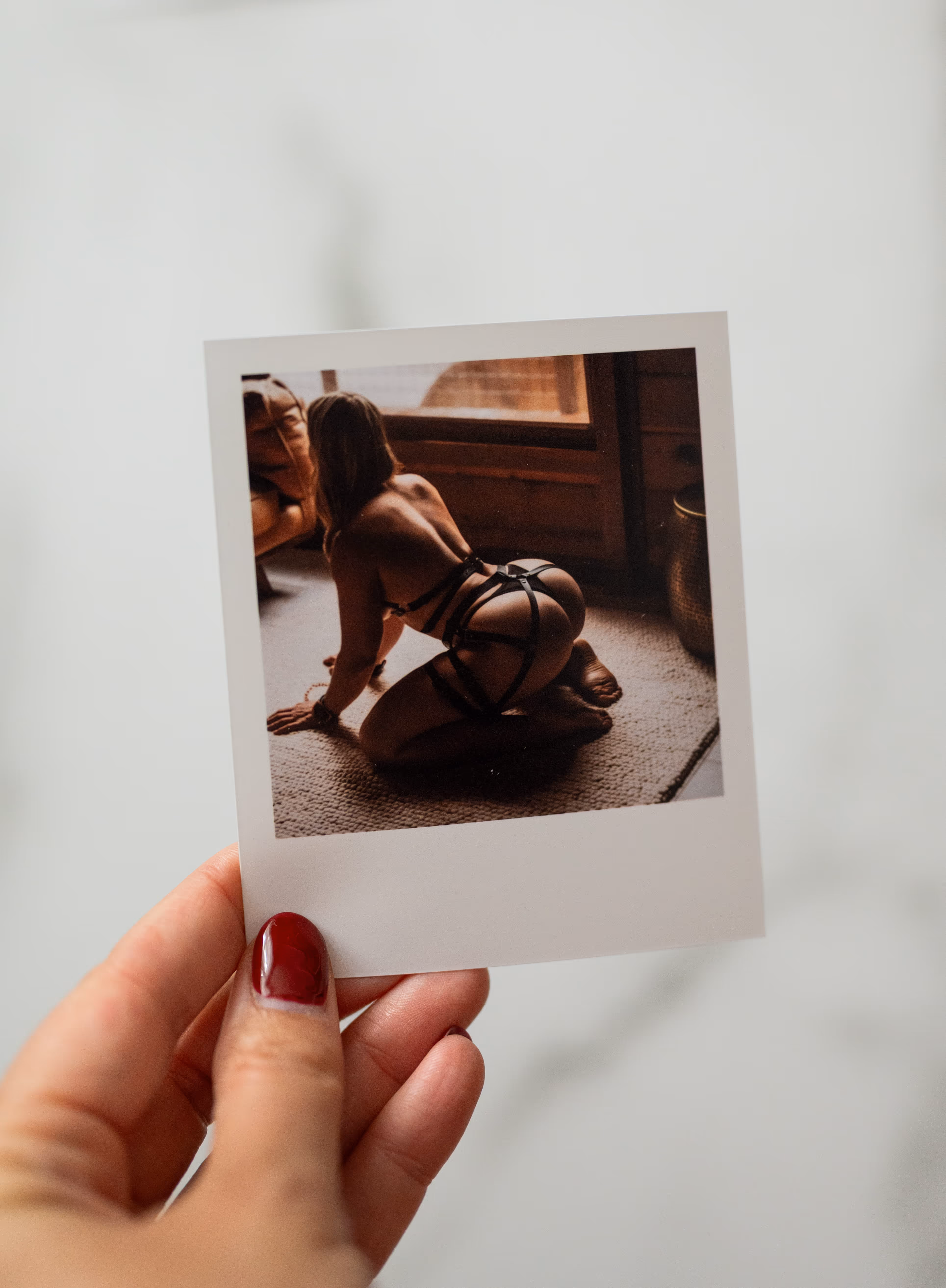 Hand with red nail polish holding a Polaroid photo of a person kneeling on carpet wearing black strappy lingerie.