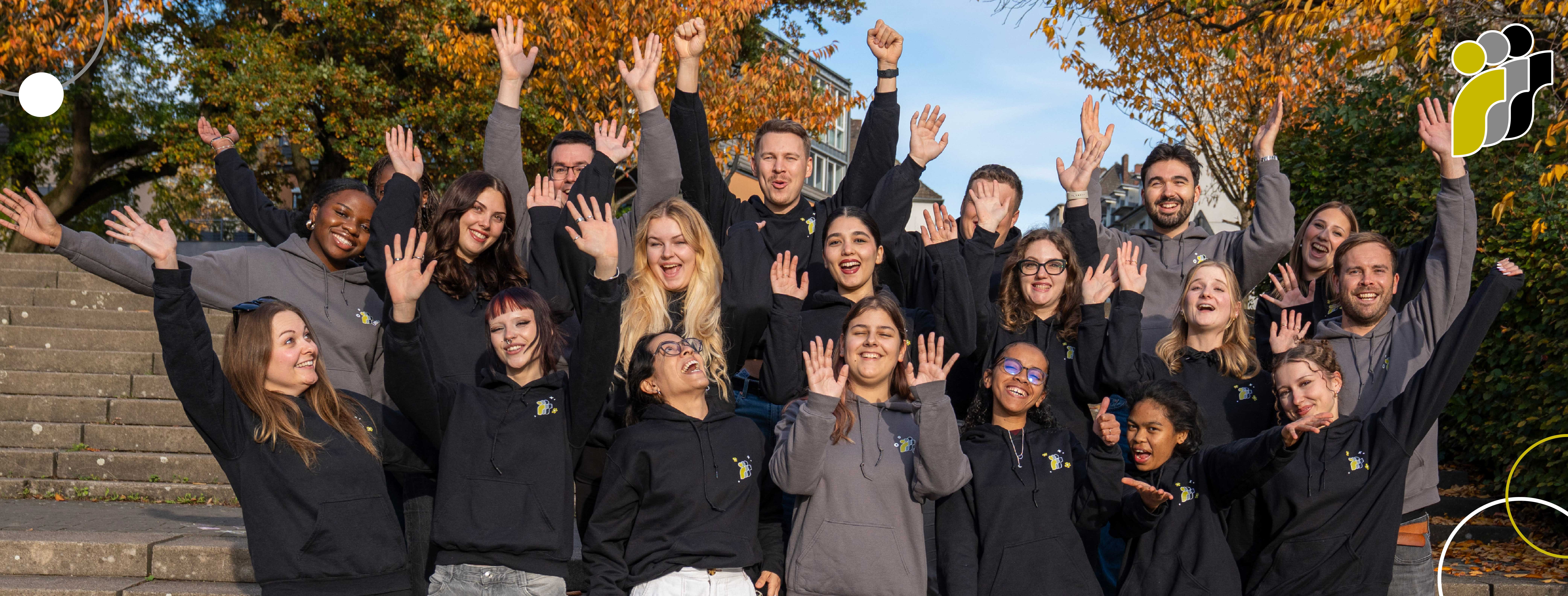 Gruppenfoto eines lächelnden, vielfältigen Teams, das im Freien vor herbstlichen Bäumen steht und die Hände jubelnd in die Luft hebt.