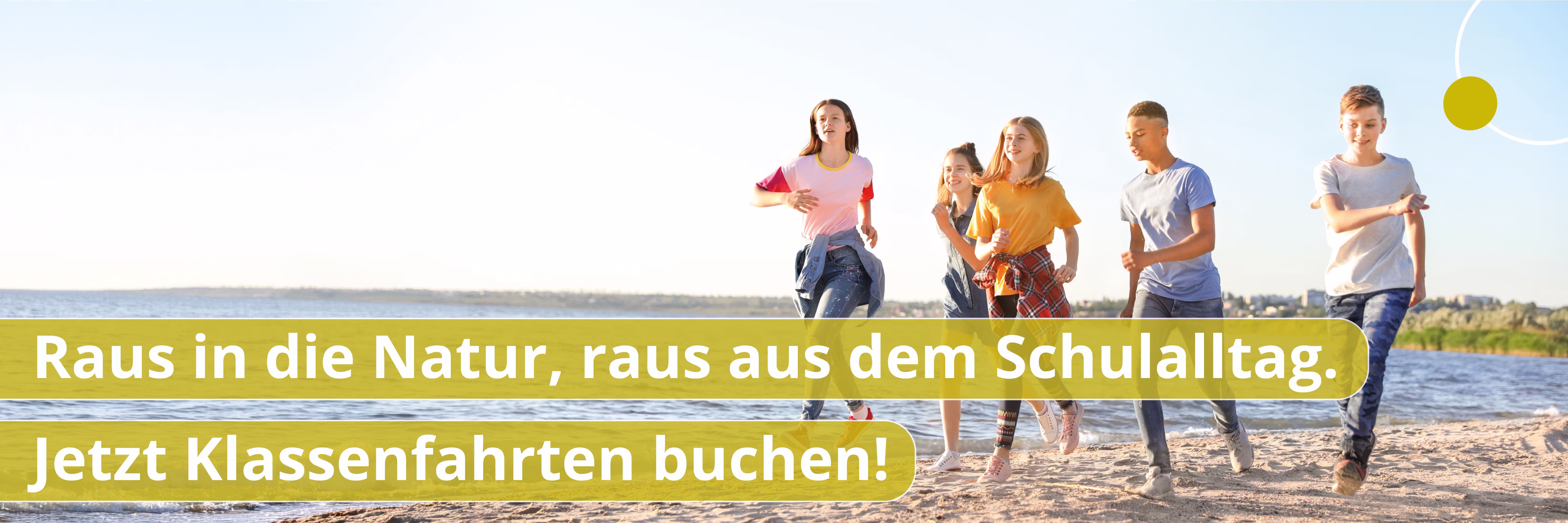Drei Kinder mit Rucksäcken sitzen am Seeufer, schauen auf das Wasser, eines zeigt nach rechts.
