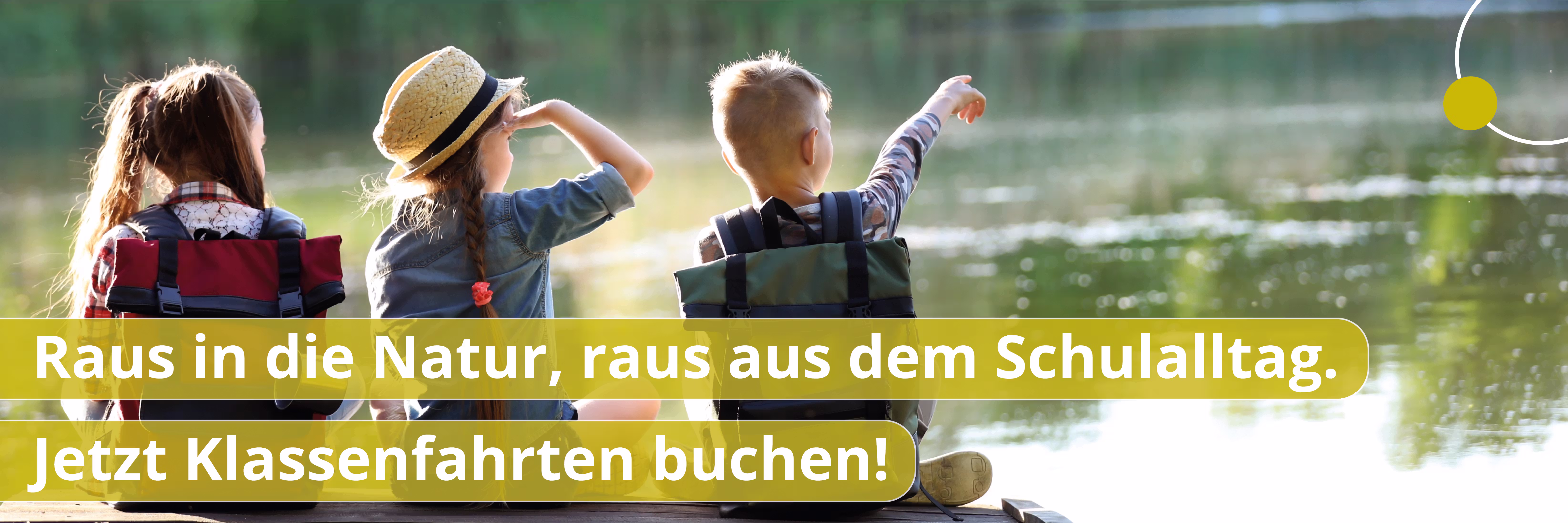 Drei Kinder mit Rucksäcken sitzen am Seeufer, schauen auf das Wasser, eines zeigt nach rechts.