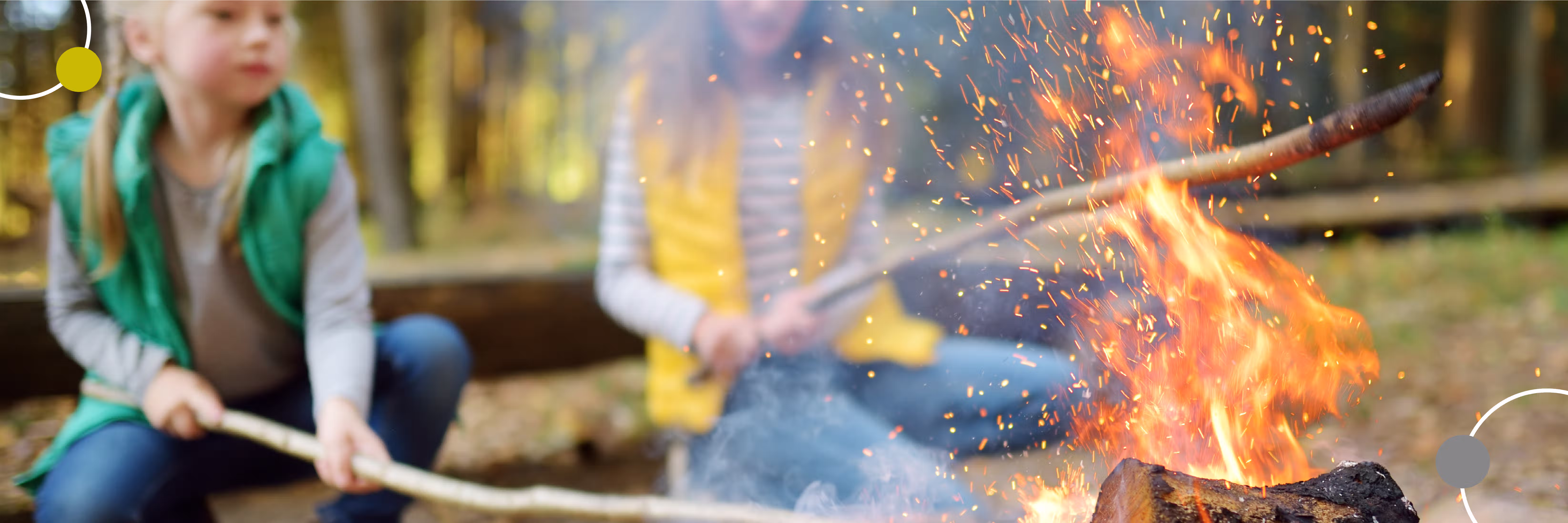 Zwei Kinder sitzen draußen am Lagerfeuer und halten Stöcke in der Hand.