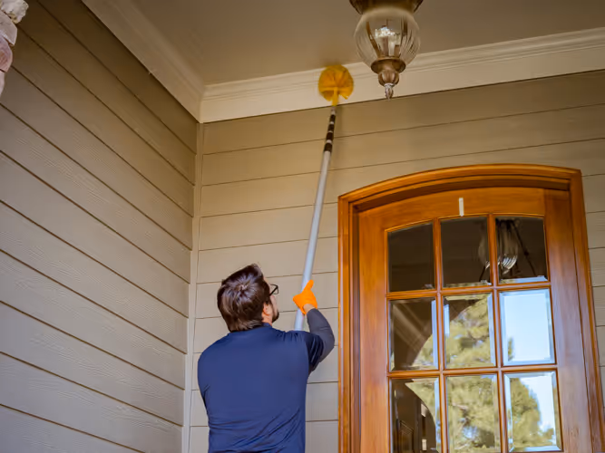 Pest Control Technician using a long-handled duster to clean the exterior of a house of cobwebs.