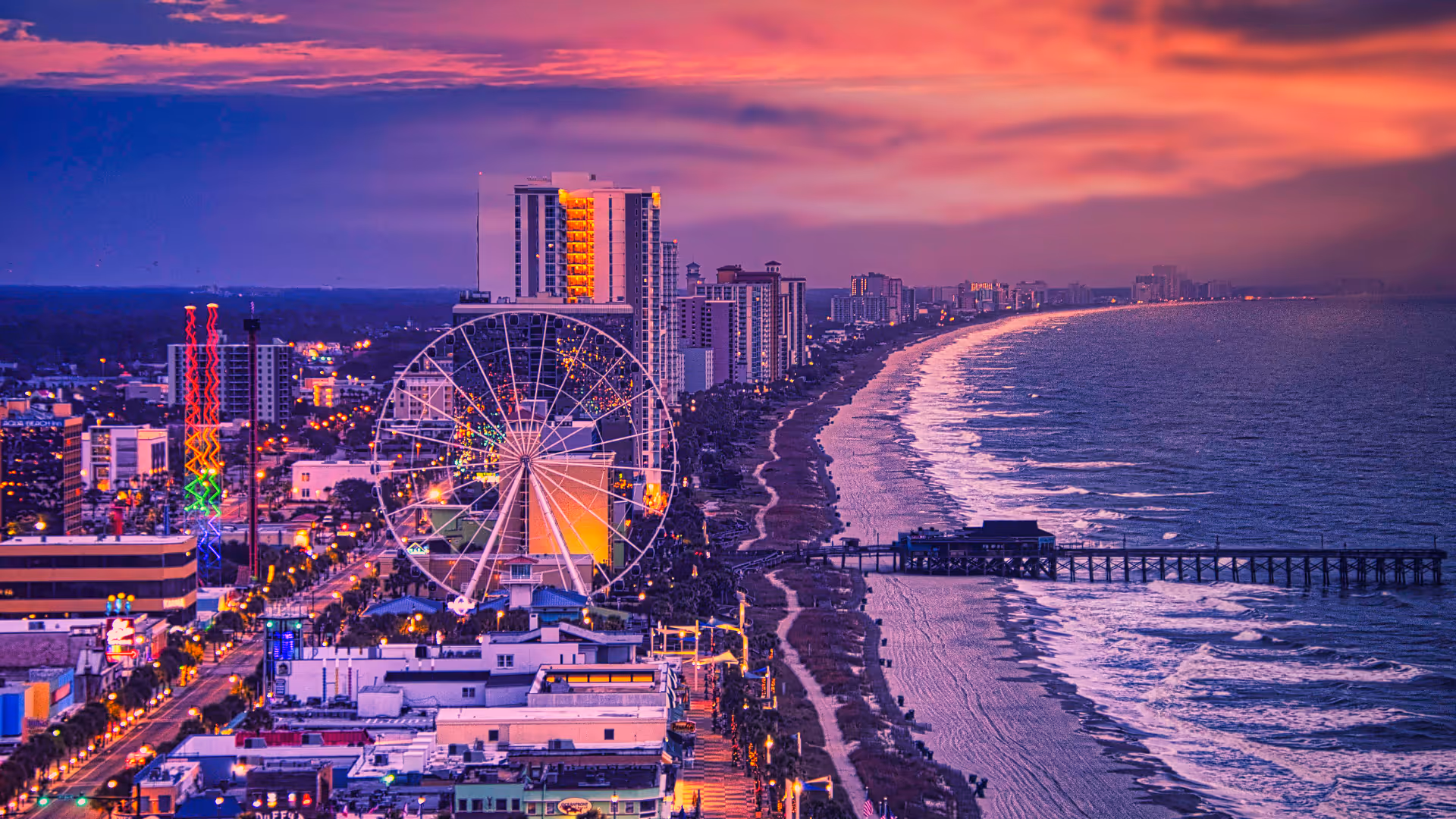Sunset view of Myrtle Beach skyline with illuminated Ferris wheel and oceanfront pier along the beach.