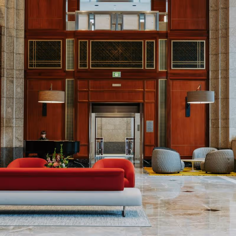 Hotel lobby with polished marble floor, modern red and gray seating, wooden panel walls, and a grand piano with a person playing.