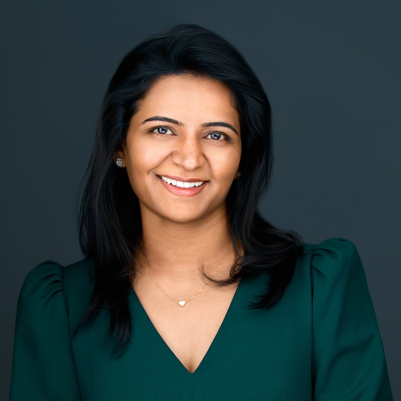 Smiling woman with dark hair wearing a green top and a heart-shaped necklace against a dark background.