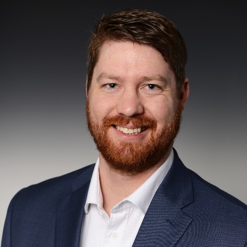Smiling man with red hair and beard wearing a navy blazer and white shirt against a gray gradient background.