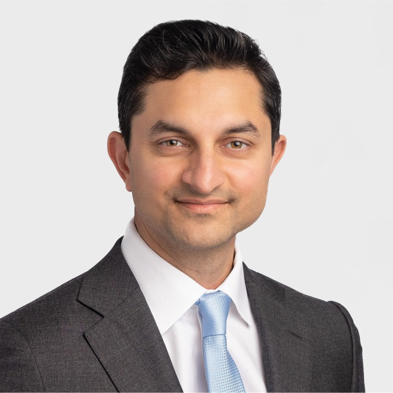 Smiling man with dark hair wearing a gray suit, white shirt, and light blue tie against a light gray background.