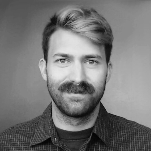 Black and white portrait of a man with short, styled hair, a thick mustache, and a septum piercing, wearing a checkered shirt.