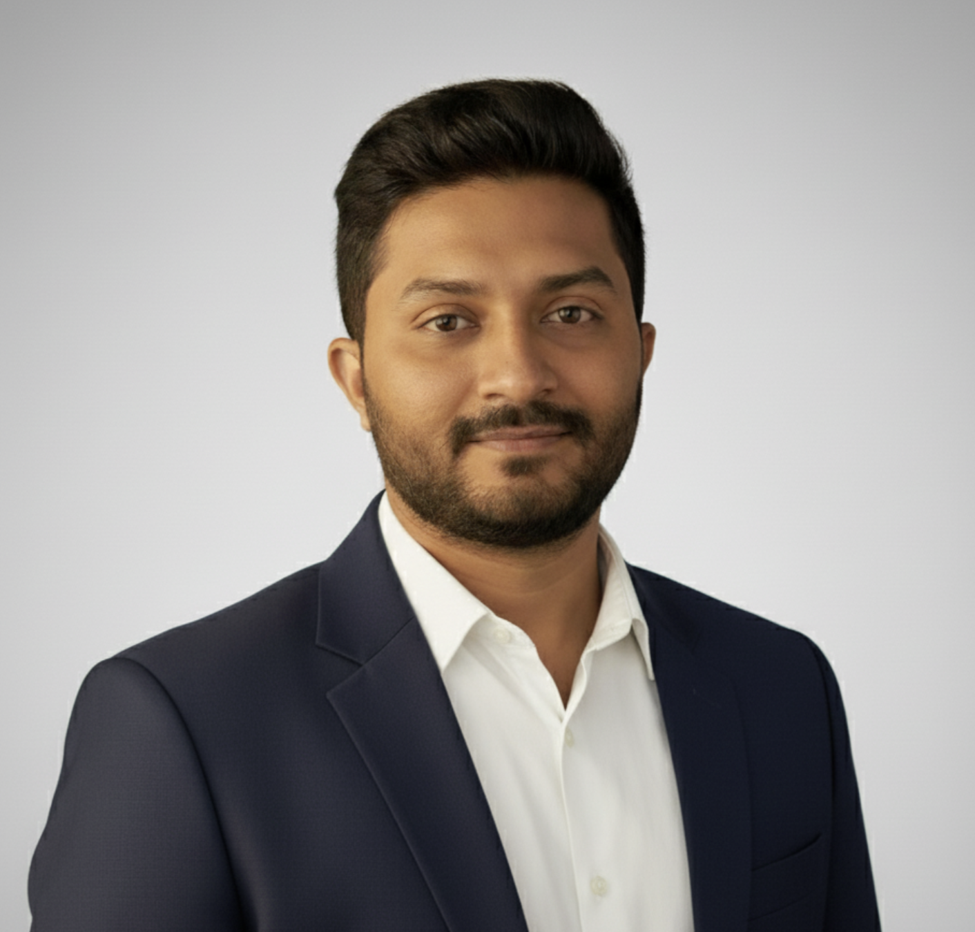 Portrait of a man with dark hair and a beard, wearing a navy blazer and white shirt against a light gray background.