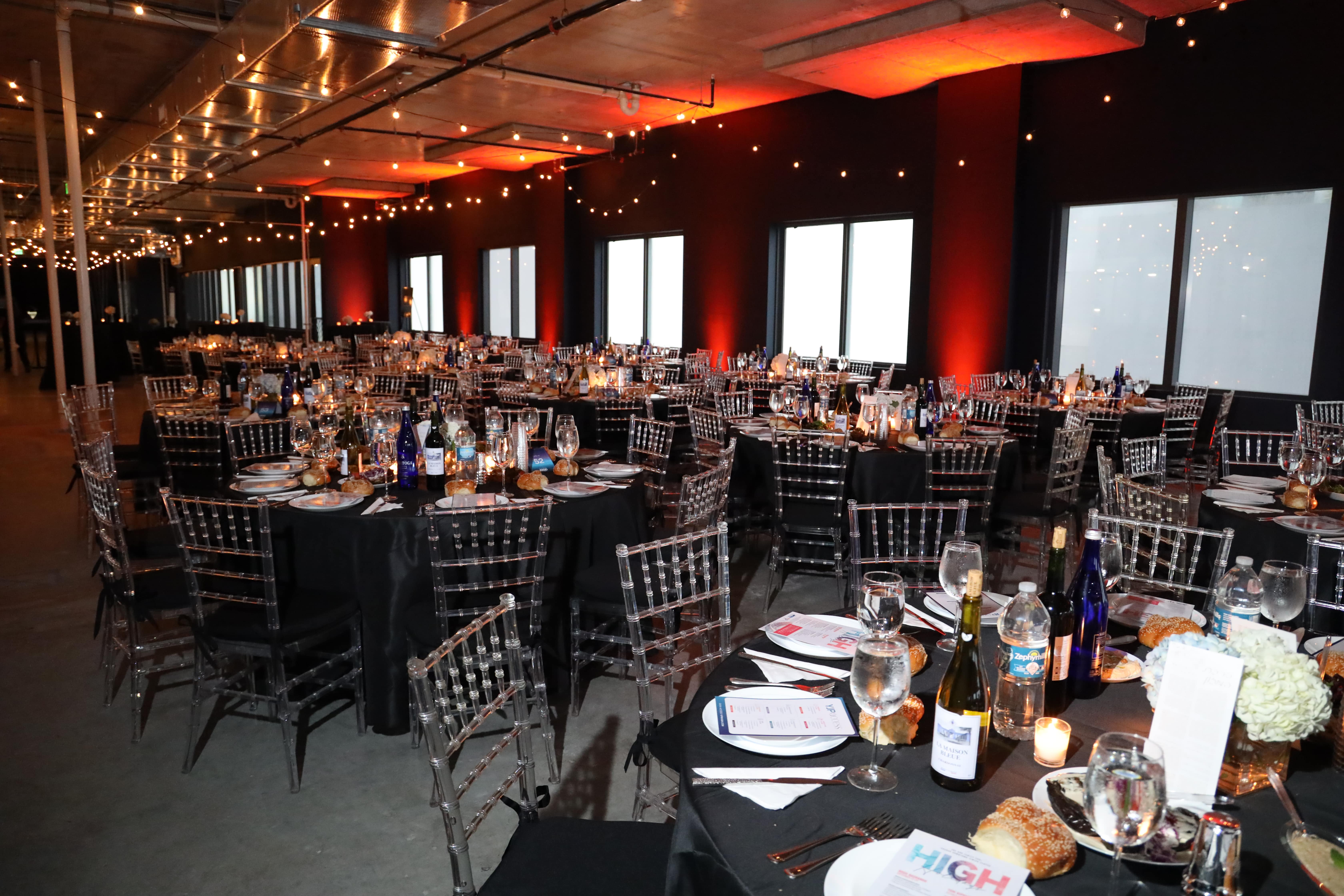 Formal evening event space featuring multiple black-draped round tables set with clear chairs, dramatic red and amber lighting on the columns, and string lights overhead.