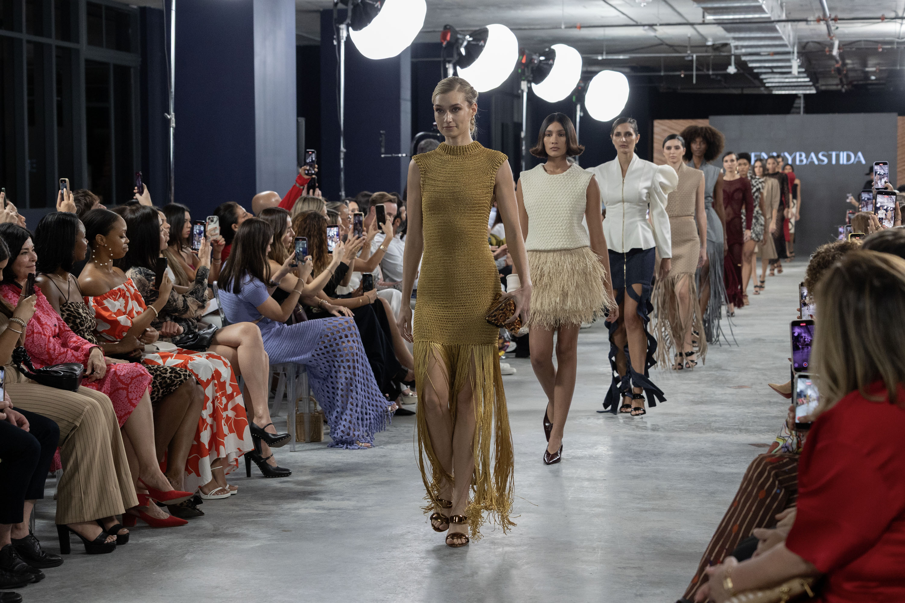 Fashion show at the Gale Miami Hotel's exhibition center, featuring a designer and models on the runway, with an audience actively taking photos.