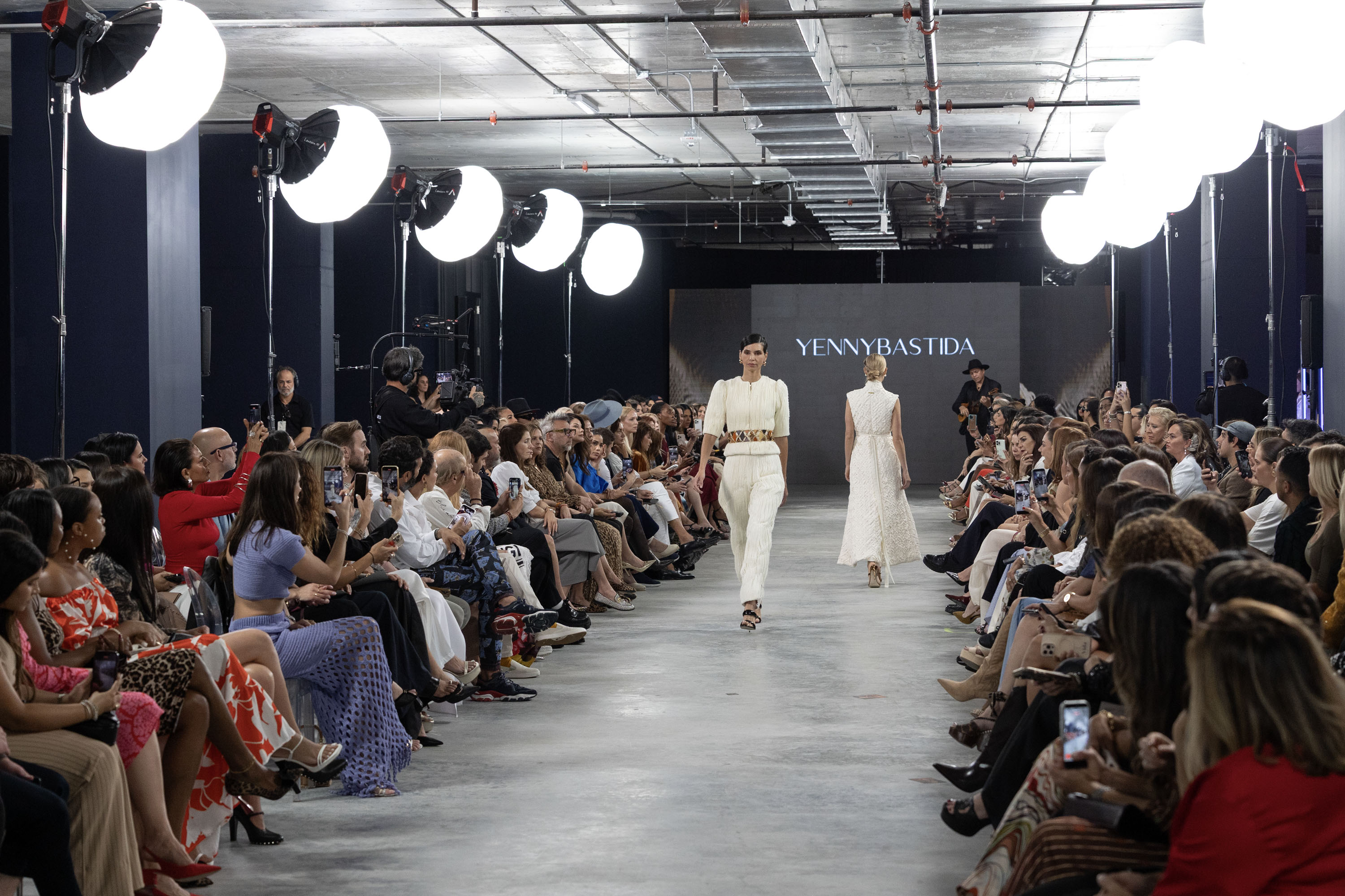 Two models walk down a fashion show runway at the Gale Miami Hotel's exhibition center, displaying white textured outfits.