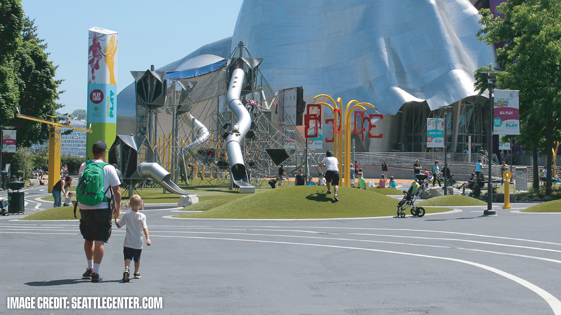 Children and adults enjoying an outdoor playground with slides and climbing nets near the metallic façade of a modern building.