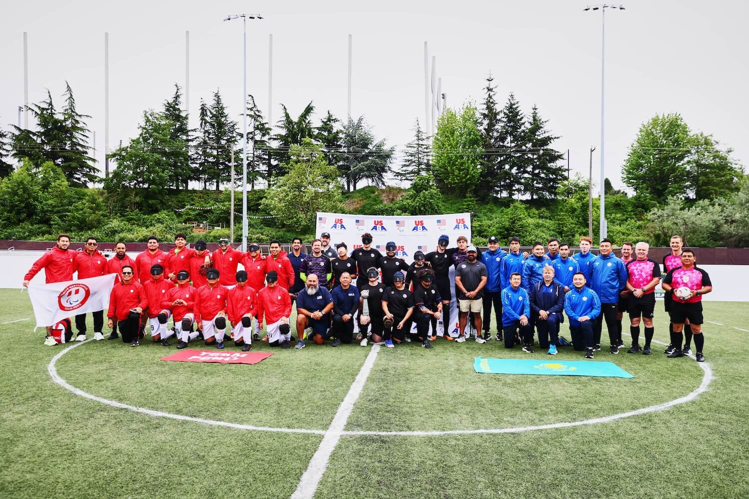Group photo of blind soccer teams posed together on an outdoor field. Team Peru stands on the left in red, Team USA in the center in black, Team Kazakhstan on the right in blue, and referees on the far right in bright pink. Flags and banners are displayed in front of the teams.