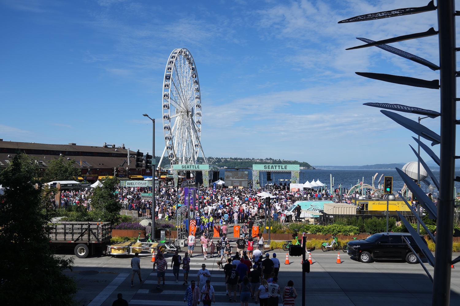 High-angle view of a large crowd gathered at Seattle’s Waterfront Park at Pier 58 under a clear blue sky, with the Seattle Great Wheel and Elliott Bay in the background during a Juneteenth Celebration and March to the Match event.