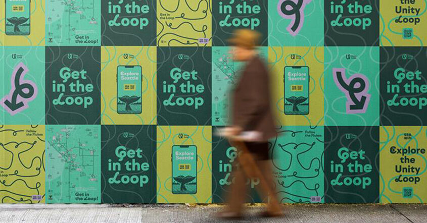 A person blurred in motion walks past a wall covered in a grid pattern of green and yellow posters advertising "Get in the Loop" and "Explore Seattle."