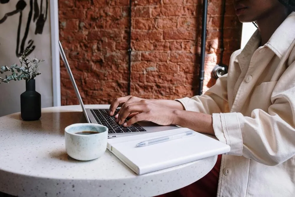 Personne travaillant sur un ordinateur portable dans un café cosy, avec une tasse de café, un carnet et un stylo posés sur la table.