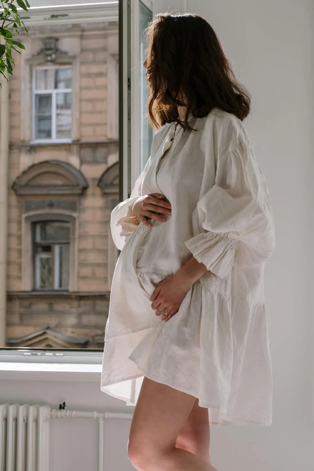 Femme enceinte en robe blanche, debout près d'une fenêtre ouverte, baignée de lumière naturelle.
