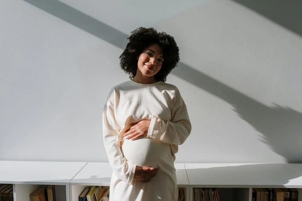 Femme enceinte souriante, en robe crème, debout dans un intérieur lumineux, les mains posées sur son ventre