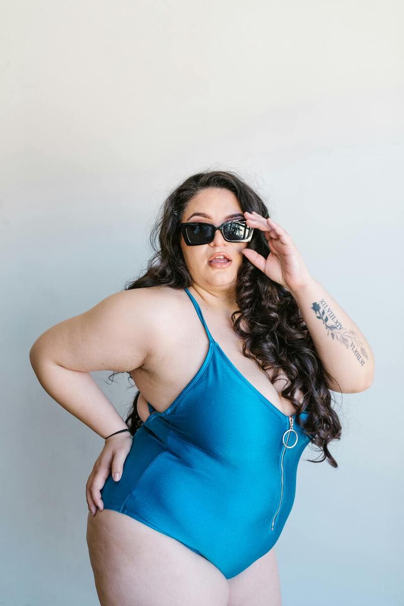 Femme confiante en maillot une pièce bleu, portant des lunettes de soleil, posant avec assurance et fierté, incarnant la beauté et la puissance de tous les corps.