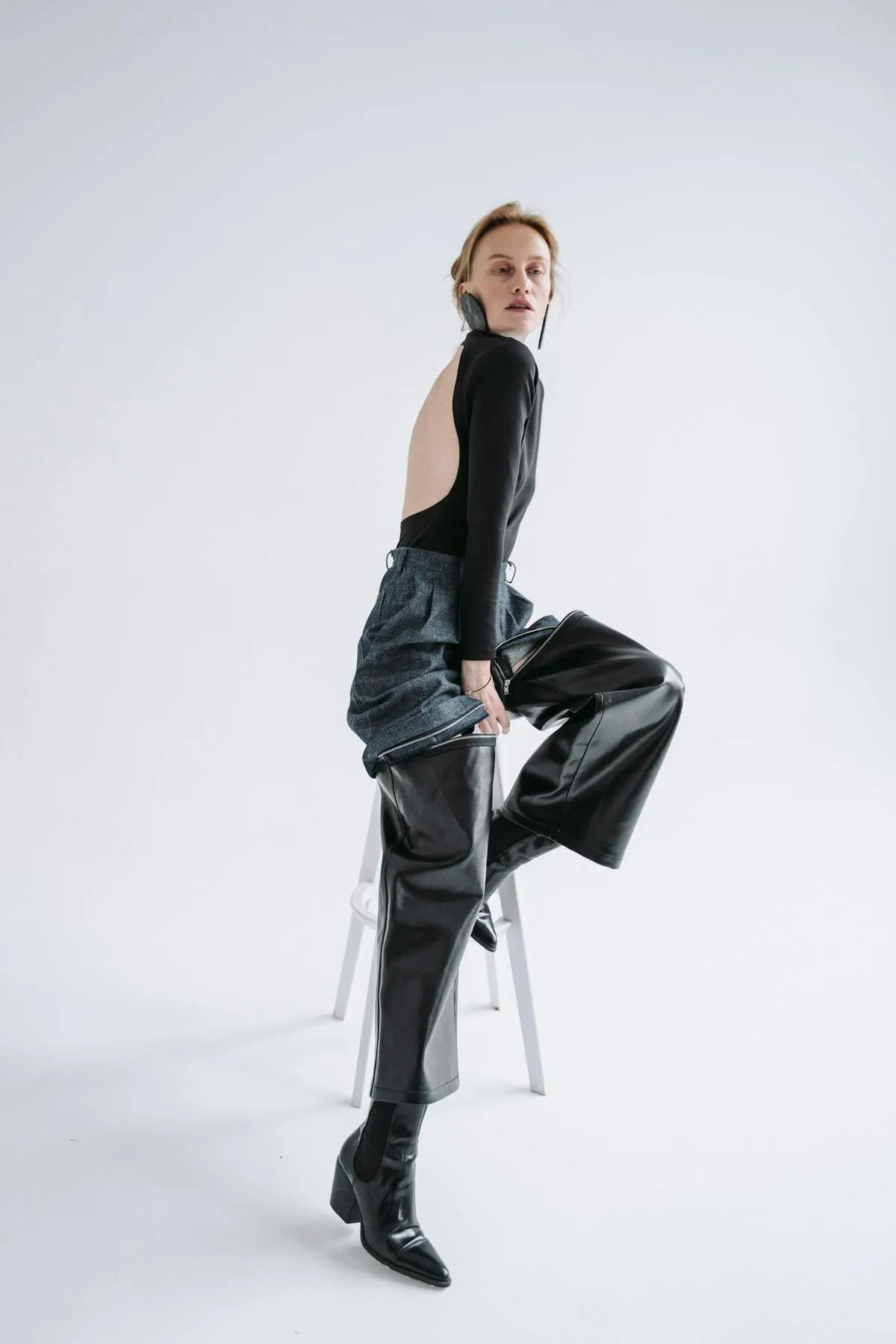 Femme de profil posant sur un tabouret, portant un body noir et un pantalon bi-matière jean et cuir avec des bottines noires, le tout donnant un look très sophistiqué