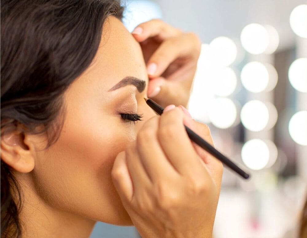 Maquilleuse appliquant un trait d'eyeliner sur une femme aux yeux fermés, en préparation d’un maquillage de fêtes