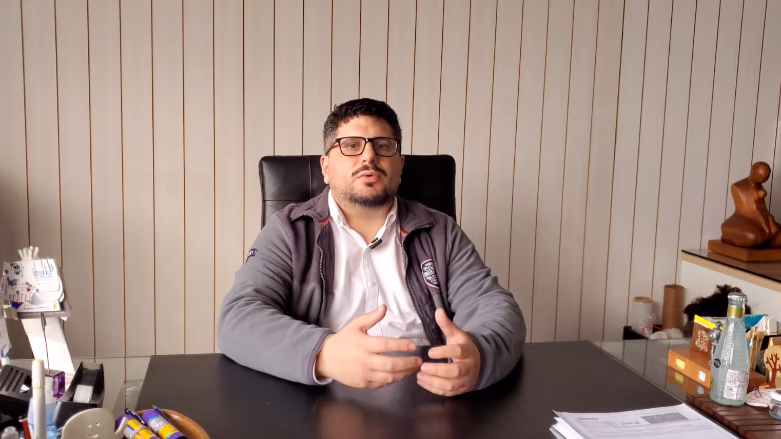 Hombre con gafas y chaqueta gris sentado en escritorio con documentos y objetos de oficina.