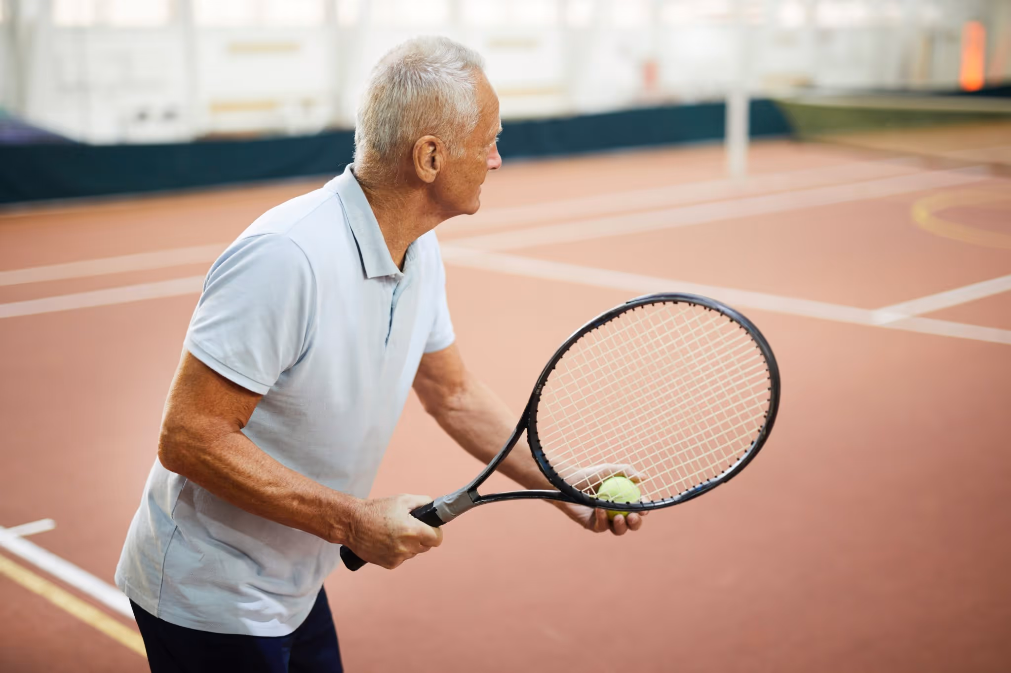 Oudere man in wit poloshirt die zich voorbereidt om tennisbal te serveren op een indoor tennisbaan.