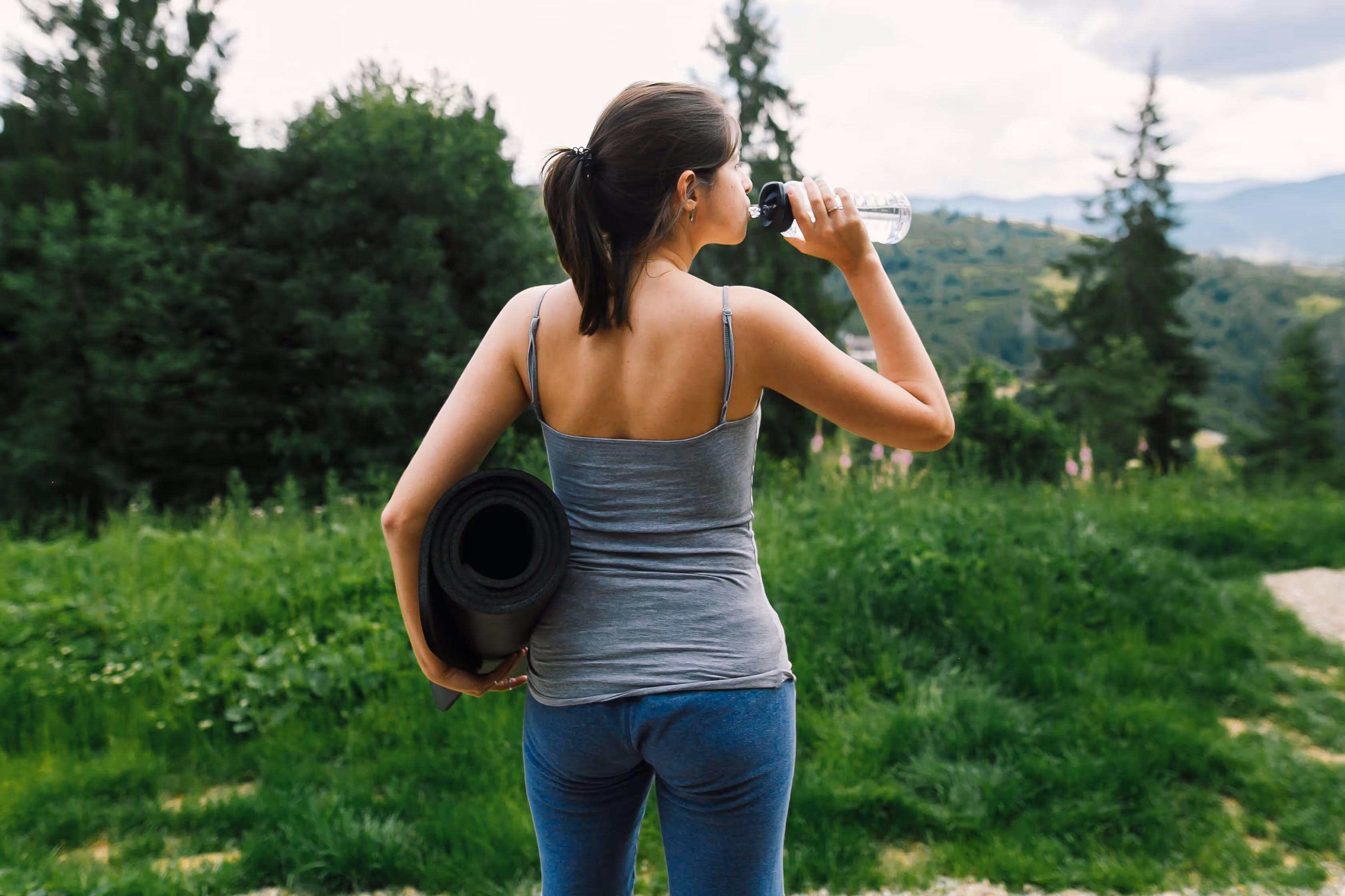 Vrouw in sportkleding die een yogamat draagt en water drinkt in een groene buitenomgeving.