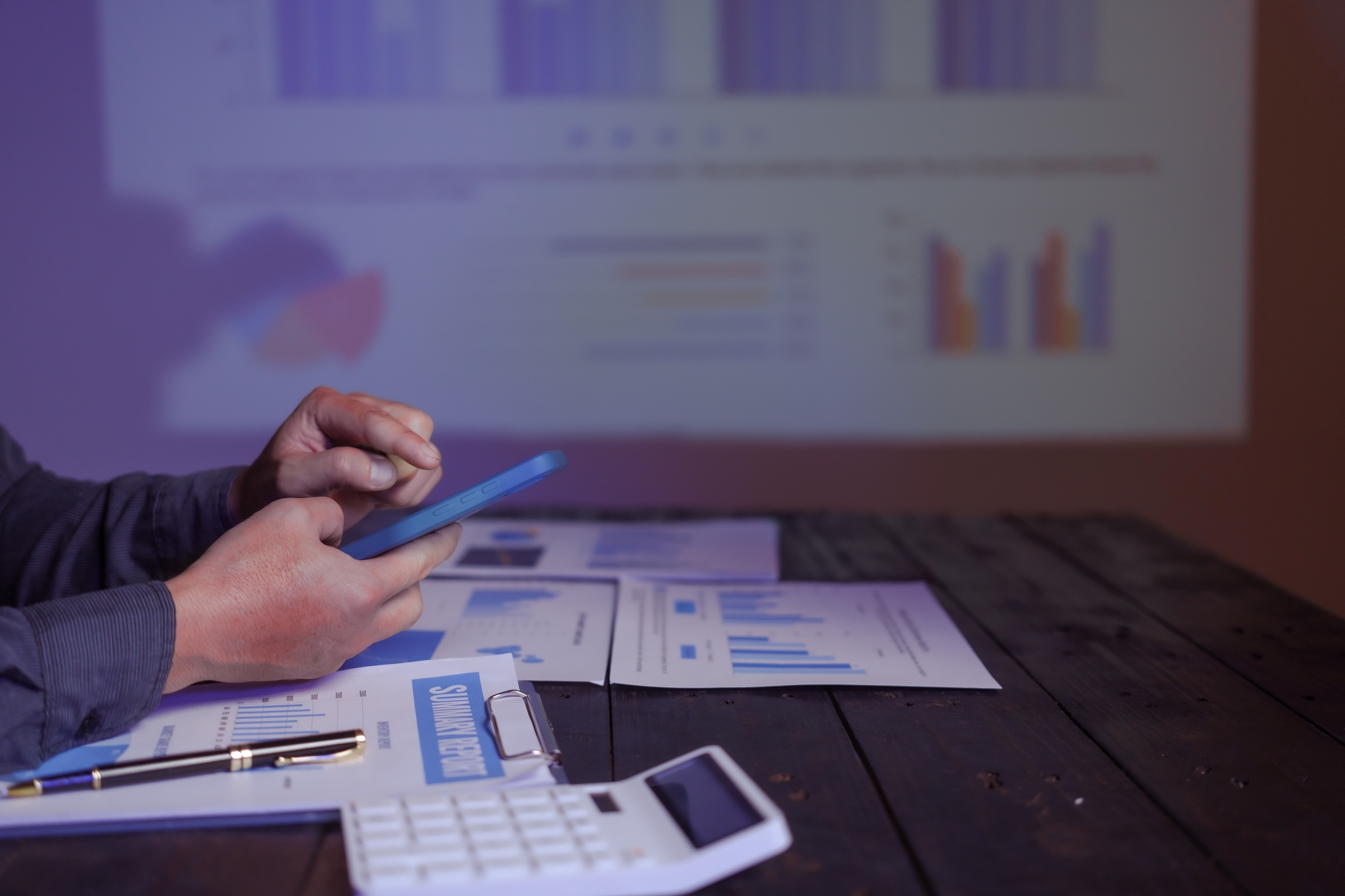 Eine Nahaufnahme zeigt die Hände einer Person, die ein blaues Smartphone bedient. Die Person sitzt an einem dunklen Holztisch, auf dem ausgedruckte Berichte mit Diagrammen, ein Taschenrechner und ein Stift liegen. Im unscharfen Hintergrund wird eine Präsen