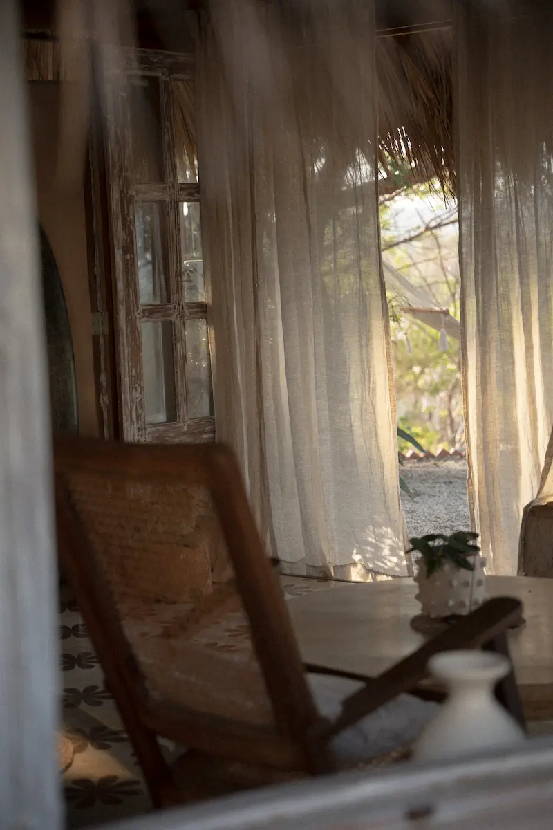 A view through the window to a chair and curtains blowing in the wind.